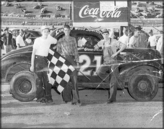 Three men standing in front of an old racing car holding a checkered flag.