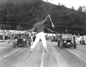 A man jumping to signal two old cars to begin racing.