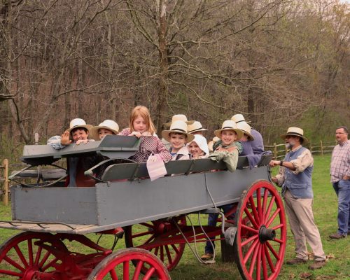 A group of children in old-fashioned clothing seated in the back of an old wagon.