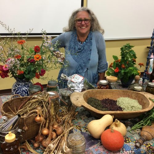 A woman standing behind a table holding a variety of herbs and gourds.