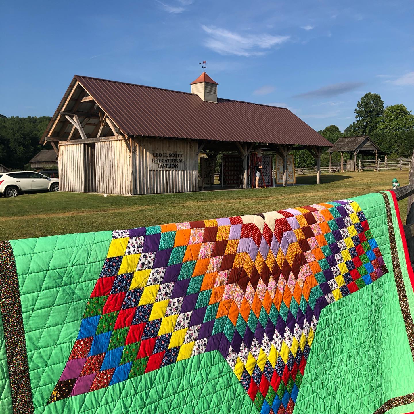 A quilt draped over a fence with a wooden building in the background.