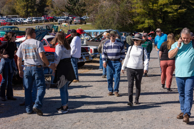 People walking past a display of antique cars and trucks along a gravel road.